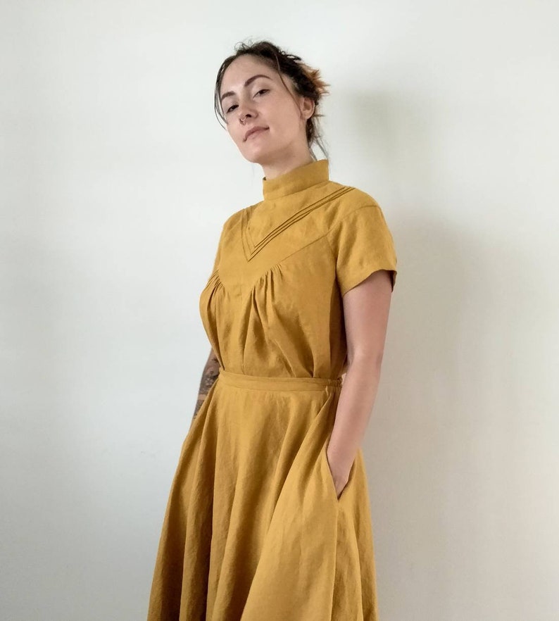 May include: A mustard yellow linen dress with a high neck and short sleeves. The dress has a gathered bodice and a full skirt with pockets.
