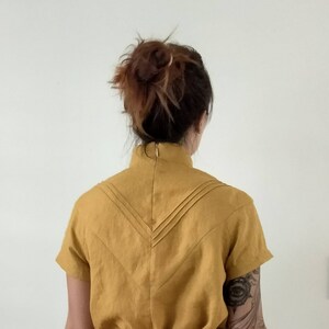 May include: A mustard yellow linen dress with a high neckline and short sleeves. The back of the dress features a V-shaped design with pleats.