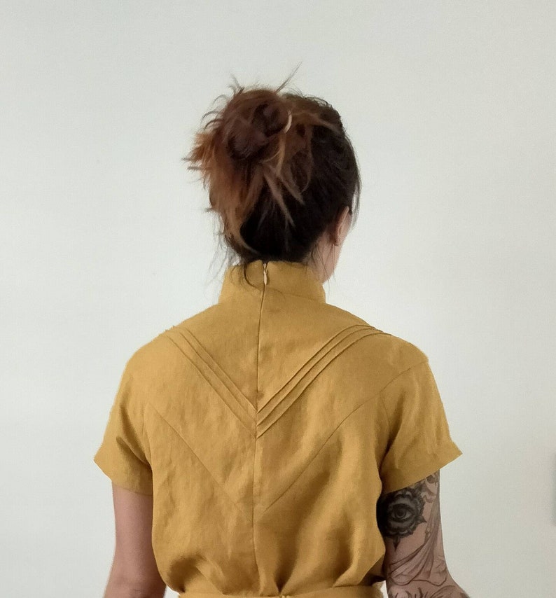 May include: A mustard yellow linen dress with a high neckline and short sleeves. The back of the dress features a V-shaped design with pleats.