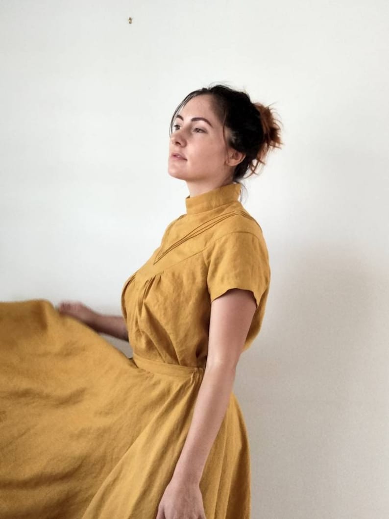 May include: A woman wearing a mustard yellow linen dress with a high neck and short sleeves. The dress has a gathered skirt and a tie belt.