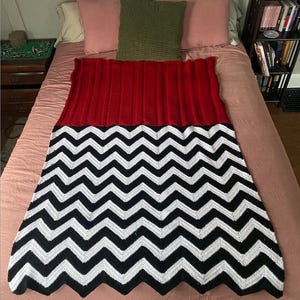 May include: A red and black crocheted blanket with a chevron pattern. The blanket is on a pink bed with two pillows.