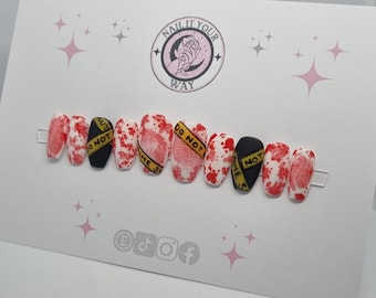 May include: A set of press-on nails with a crime scene theme. The nails feature a white base with red blood splatter and fingerprint designs. Some nails have black bases with yellow "DO NOT CROSS CRIME SCENE" tape.