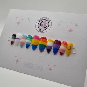 May include: A set of ten artificial nails, each painted with a different LGBTQ+ pride flag design. The nails are almond-shaped and displayed on a white card with the text "NAIL IT YOUR WAY" and social media icons.