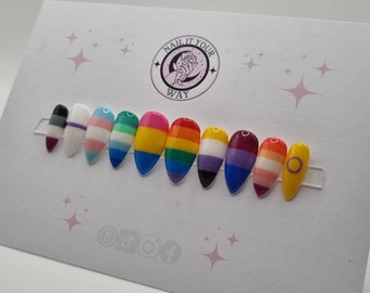 May include: A set of ten artificial nails, each painted with a different LGBTQ+ pride flag design. The nails are almond-shaped and displayed on a white card with the text "NAIL IT YOUR WAY" and social media icons.