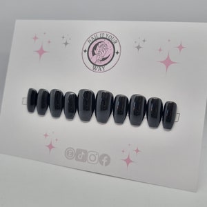 Puede incluir: Un juego de diez uñas postizas negras brillantes dispuestas sobre una tarjeta blanca. La tarjeta presenta un logotipo circular con el texto "NAIL IT YOUR WAY" e iconos de redes sociales. Diseños de estrellas rosas están dispersos en la tarjeta.