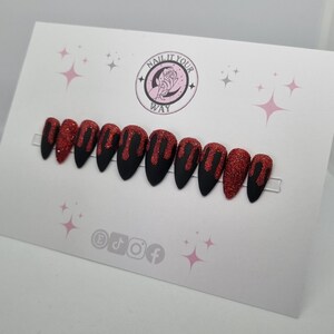 May include: A set of stiletto-shaped false nails. The nails are black with a red glitter design that resembles dripping blood. The nails are displayed on a white card with the text "NAIL IT YOUR WAY" and social media icons.