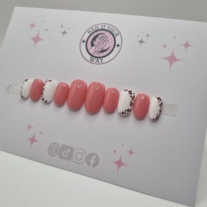May include: A set of oval-shaped press-on nails. The nails alternate between solid coral pink and white with black leopard print tips. The card behind the nails has the text "NAIL IT YOUR WAY" and social media icons.