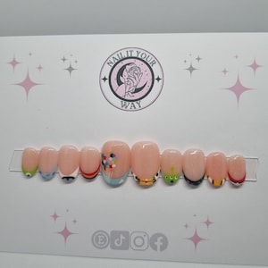 Può includere: Un set di dieci unghie finte con una base rosa chiaro e vari disegni a tema cartoni animati. I disegni includono occhi, palloncini e un alieno verde. Le unghie sono esposte su una carta bianca con la scritta "NAIL IT YOUR WAY".
