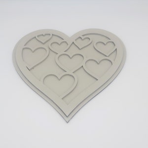 Puede incluir: Una decoración de pared de madera en forma de corazón de color gris claro con un diseño en capas que presenta varios corazones recortados.