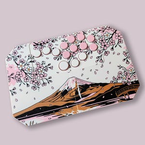 May include: A rectangular arcade controller featuring a white base with a graphic print of a mountain, a bridge, and pink cherry blossoms. The top surface includes a cluster of eleven circular buttons in alternating shades of white and light pink.