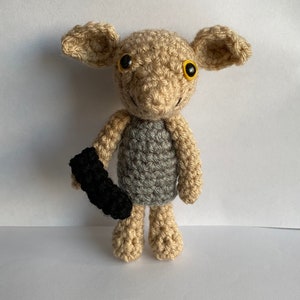May include: A crocheted doll in the shape of a creature with large ears and yellow eyes. The doll is primarily beige with a gray torso and a black object in its hand. The doll is handmade and has a whimsical appearance.