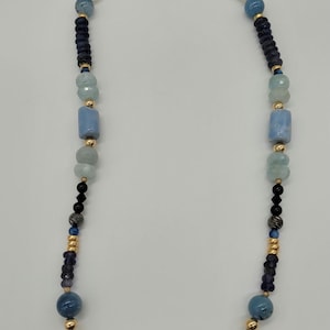 May include: A necklace featuring a variety of blue and gray beads, including round, cylindrical, and irregular shapes. The beads are strung on a gold-colored wire, with a gold clasp. The necklace is displayed against a white background.
