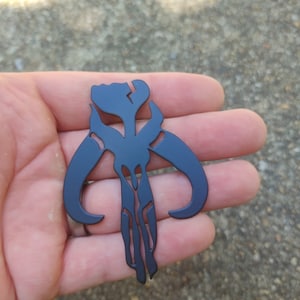 May include: A black metal Mandalorian symbol, also known as the "Death Watch" symbol, is shown in a hand. The symbol is a stylized skull with two horns.