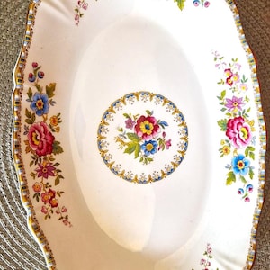 May include: A white oval serving dish with a scalloped edge and a floral design. The flowers are pink, red, blue, and yellow. The dish has a gold trim.