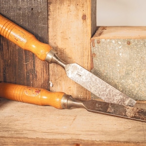 Può includere: Due scalpelli per legno con manici in legno marrone chiaro e ghiere in metallo. Le lame in acciaio mostrano segni di invecchiamento, ossidazione e usura. Uno scalpello è posizionato diagonalmente sull'altro, entrambi con steli metallici piatti.