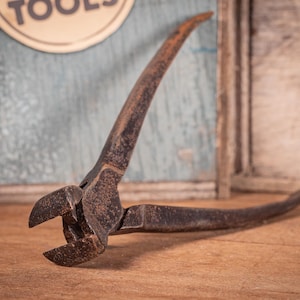 May include: Close-up of an antique, rusty metal tool. The tool has curved handles and a cutting head. The tool is likely used for cutting or shaping materials. The background includes a wooden surface and a sign that says "TOOLS".