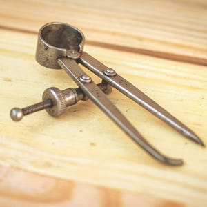 May include: A vintage metal spring-joint divider tool featuring two pointed legs connected by a circular pivot head and an adjustable threaded screw mechanism with a knurled nut for precise measurement settings and secure positioning.