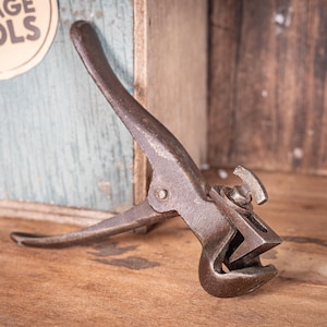 May include: A weathered metal tool, showing signs of age and rust. It features a lever-style design with a curved jaw and a pivoting joint. The background includes a wooden surface and a sign that says "VINTAGE TOOLS".