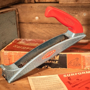 May include: A Surform plane tool featuring a bright red plastic handle and a textured, metallic gray body with a perforated blade. The tool is positioned near its original cardboard packaging box and a printed instruction sheet, both labeled Surform.