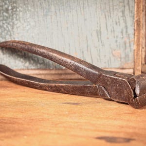 May include: A pair of weathered, dark brown metal end cutting pliers with a textured, oxidized surface. The tool features long, curved handles and a rounded, heavy-duty head designed for gripping and cutting wire or nails with precision.