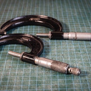 May include: Two black and silver micrometers on a green grid surface. The micrometers are used to measure the diameter of objects.