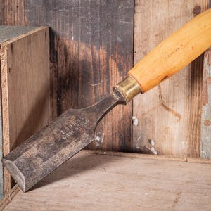 May include: A vintage wood chisel with a light-colored wooden handle and a metal blade. The blade has the text "T. SURRY CAST STEEL" etched into it. The chisel is resting on a wooden surface.