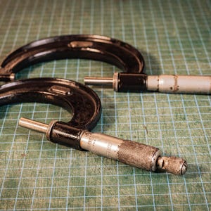 May include: Two vintage outside micrometers featuring black C-shaped frames and silver-toned metal barrels with knurled grips. These precision measuring tools display engraved measurement markings along the cylindrical sleeves and include a thimble.