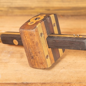 May include: A vintage wooden marking gauge with brass detailing. The tool has a dark wood body with brass inlays and a sliding head. The gauge is designed for woodworking and has a classic, well-worn appearance.