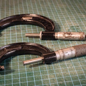 May include: Two black and silver metal outside micrometers with C-shaped frames and knurled adjustment thimbles. These precision measuring tools feature graduated scales on the barrels and spindles, showing signs of wear and metallic texture.