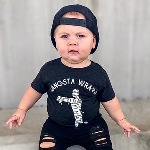 May include: A young child wearing a black t-shirt with a white mummy graphic and the text "Gangsta Wrap" is wearing black ripped jeans and a black baseball cap.