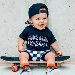 Könnte beinhalten: Ein kleines Kind trägt ein schwarz-weiß kariertes Hemd mit dem Text "Ghosting Witches" in Weiß, eine schwarze Baseballkappe und schwarz-weiß karierte Shorts und sitzt auf einem schwarzen Skateboard mit roten Rädern.