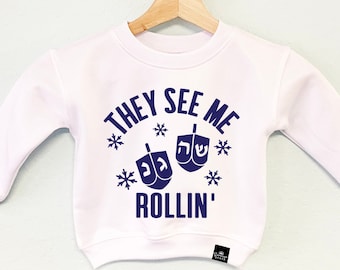 May include: White long-sleeved sweatshirt with the text "THEY SEE ME ROLLIN'" in navy blue, along with dreidel and snowflake graphics. The sweatshirt is hanging on a wooden hanger.