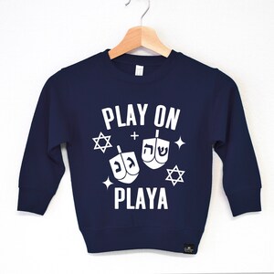 May include: A navy blue sweatshirt with white text that reads "PLAY ON + PLAYA" with a graphic of two dreidels and stars.