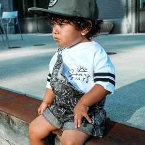 May include: A young child wearing a dark green baseball cap with a circular patch, a white t-shirt with black stripes and the text "Sonny State", and denim overalls. The child is sitting on a wooden ledge.