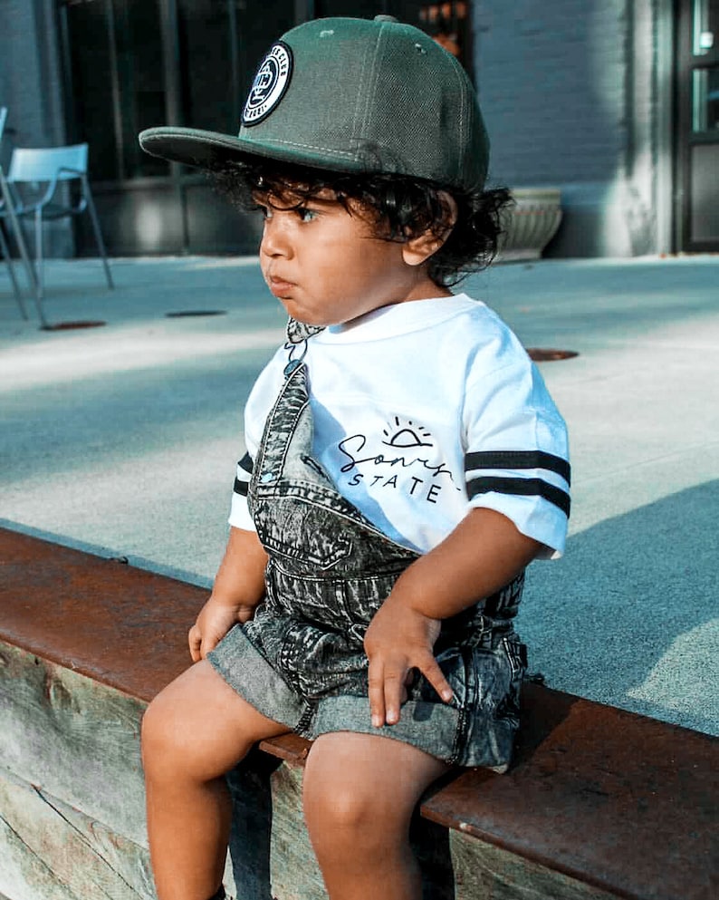 May include: A young child wearing a dark green baseball cap with a circular patch, a white t-shirt with black stripes and the text "Sonny State", and denim overalls. The child is sitting on a wooden ledge.