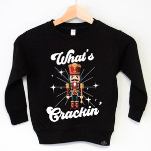 Op de afbeelding: Zwart sweatshirt met een witte afbeelding van een notenkraker en de tekst "What's Crackin'".