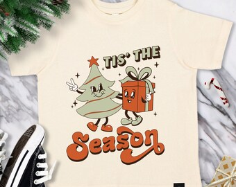 May include: Cream-coloured t-shirt with a retro Christmas design. The graphic features a cartoon Christmas tree giving a peace sign and a gift box. The text "Tis the Season" is in a retro font. A fun holiday tee.