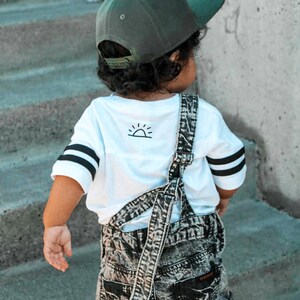 May include: A child wearing a green baseball cap, white t-shirt with black stripes on the sleeves, and denim overalls. The t-shirt has a small sun graphic on the back. The overalls are acid wash denim.