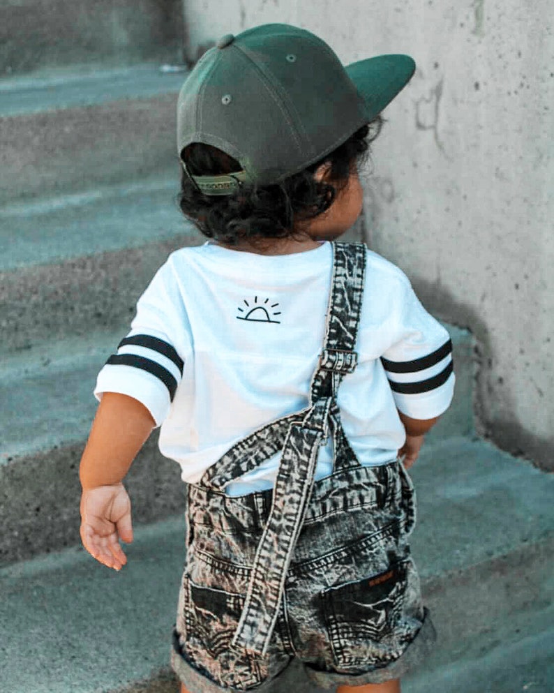 May include: A child wearing a green baseball cap, white t-shirt with black stripes on the sleeves, and denim overalls. The t-shirt has a small sun graphic on the back. The overalls are acid wash denim.