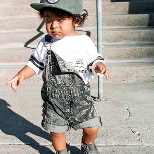 May include: A young child wearing a green baseball cap with a circular logo, a white long-sleeved shirt with black stripes and text, and denim overalls. The child is also wearing gray high-top sneakers.