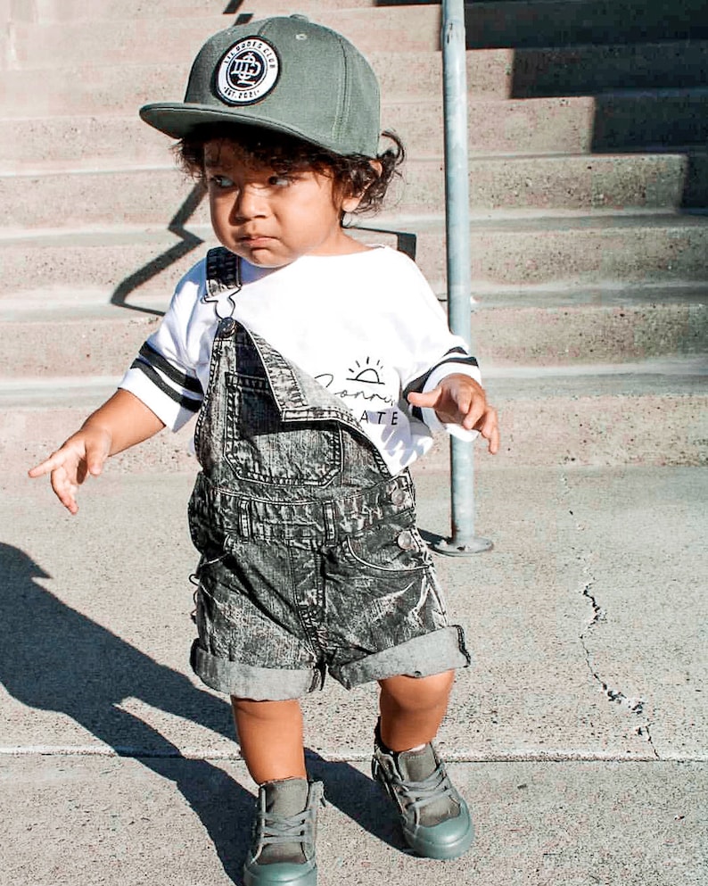 May include: A young child wearing a green baseball cap with a circular logo, a white long-sleeved shirt with black stripes and text, and denim overalls. The child is also wearing gray high-top sneakers.
