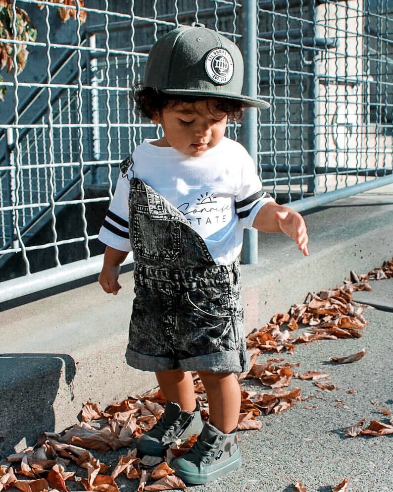 May include: A young child wearing a gray baseball cap with a circular patch, a white t-shirt with "Sunrise State" text, and denim overalls. The child is also wearing gray high-top sneakers. The image is taken outdoors.