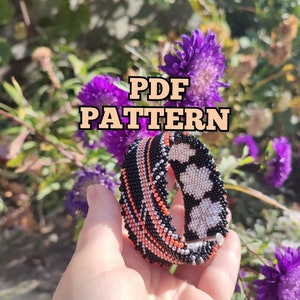 May include: A black, silver, and orange beaded bracelet with a geometric pattern. The bracelet is held in a hand against a background of purple flowers. The text "PDF PATTERN" is displayed above the bracelet.