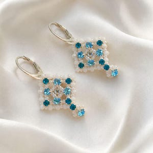 May include: A pair of dangle earrings featuring a square-shaped beaded design. The earrings are crafted with small white beads and accented with light blue and dark teal round crystals. Each earring is attached to a silver-toned metal leverback.