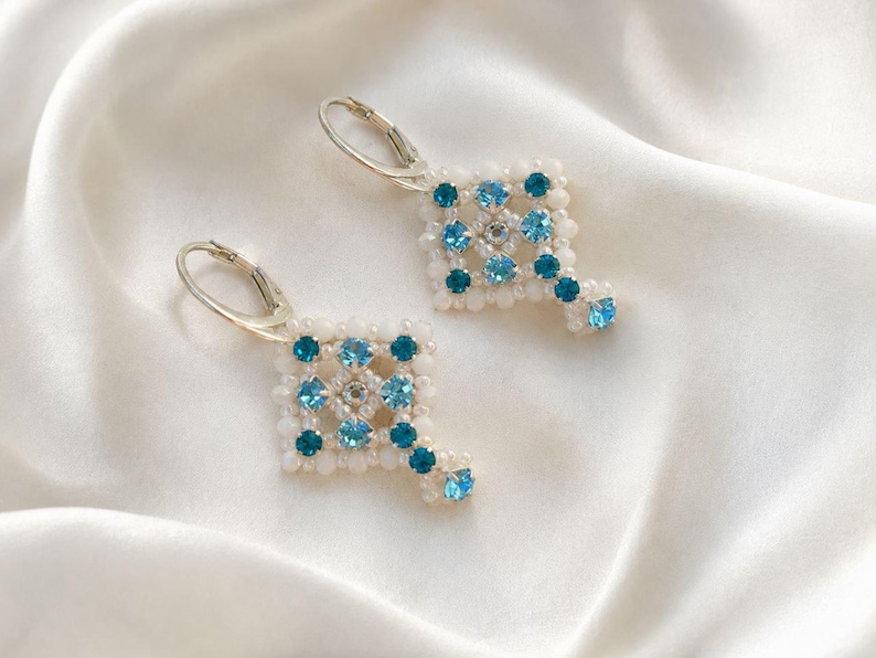 May include: A pair of dangle earrings featuring a square-shaped beaded design. The earrings are crafted with small white beads and accented with light blue and dark teal round crystals. Each earring is attached to a silver-toned metal leverback.