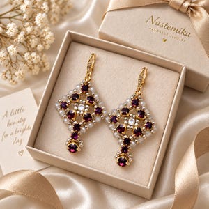 May include: A pair of gold-toned dangle earrings featuring a diamond-shaped lattice design with deep purple and clear crystals. The earrings are presented in a beige gift box labeled Nastemika with a card reading A little beauty for a brighter day.