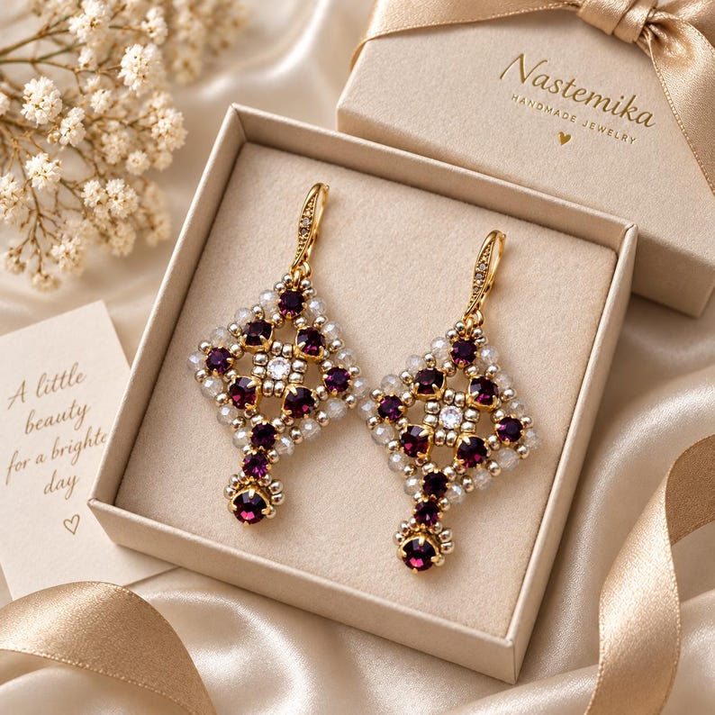 May include: A pair of gold-toned dangle earrings featuring a diamond-shaped lattice design with deep purple and clear crystals. The earrings are presented in a beige gift box labeled Nastemika with a card reading A little beauty for a brighter day.
