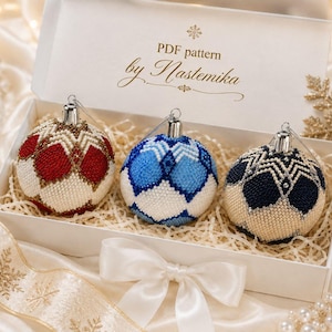 May include: A white gift box containing three spherical ornaments decorated with intricate beadwork in red, blue, and dark blue diamond patterns. The box lid features the text "PDF pattern by Nastemika" printed in gold script with a snowflake icon.