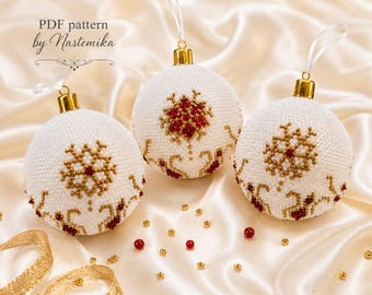 Op de afbeelding: Drie bolvormige kerstornamenten met witte, met kralen bewerkte oppervlakken en gouden en rode sneeuwvlokpatronen. Elk ornament heeft een goudkleurige dop en een wit ophanglusje. De tekst "PDF pattern by Nastemika" is zichtbaar in de linkerbovenhoek.