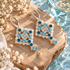 May include: A pair of dangle earrings featuring a square geometric pattern crafted from small white beads and alternating light and dark blue crystals. Each earring is finished with a silver-toned leverback closure and a single blue crystal drop.